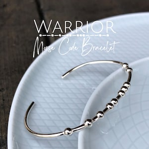 Peut inclure: Bracelet argenté avec une rangée de petites perles épellant le mot "WARRIOR" en code Morse.