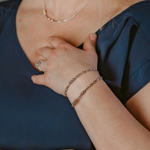 May include: Two silver beaded bracelets stacked on a person's wrist. The bracelets are made of small, evenly spaced beads.