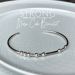 May include: A silver bangle bracelet with a series of small beads. The bracelet is on a white plate and the text "STRONG Morse Code Bracelet" is above it.