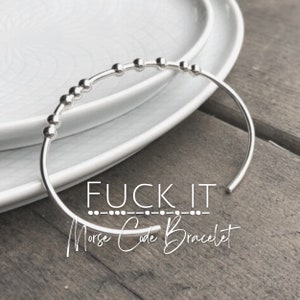Peut inclure: Bracelet argenté avec de petites perles qui épellent "FUCK IT" en code Morse. Le bracelet est sur une assiette blanche et une surface en bois. "Bracelet en code Morse" est écrit sous le bracelet.