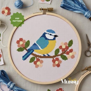 Op de afbeelding: Een kruissteekpatroon van een blauwe, gele en witte vogel die op een bloeiende tak zit, geplaatst in een houten borduurring. Het ontwerp bevat roze bloemen en groene bladeren. De woorden "PDF PATTERN" staan in een groene cirkel.