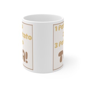 Puede incluir: Taza de cerámica blanca con un diseño rectangular beige con el texto "1 Potato, 2 Potato, 3 Potato, TA!" en letras marrones y doradas. La taza tiene un asa estándar y un acabado liso y brillante.