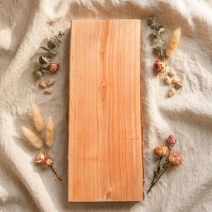 Può includere: Un tagliere rettangolare in legno con bordo naturale, in una calda tonalità marrone chiaro. La tavola è decorata con fiori secchi, piccole pietre e rametti di verde, creando un'estetica rustica e naturale.