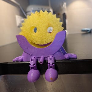 May include: A yellow, circular, serrated-edge sponge with a smiley face cutout, held by a purple, 3D-printed plastic holder featuring articulated legs and arms that allow the figure to sit on the edge of a sink.