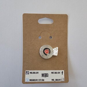 Puede incluir: Un pin de esmalte plateado con un diseño de disco de vinilo con centro rojo y un pequeño tocadiscos. El pin está sujeto a una tarjeta marrón con la palabra "MERCI" e iconos de redes sociales.