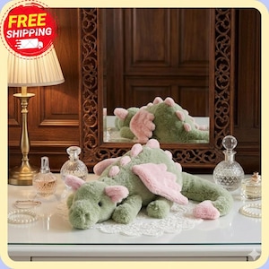 Op de afbeelding: Een groene pluche draak met roze vleugels en accenten ligt op een wit oppervlak. De weerspiegeling van de draak is te zien in een spiegel erachter. De sc&egrave;ne omvat een lamp, parfumflesjes en sieraden.