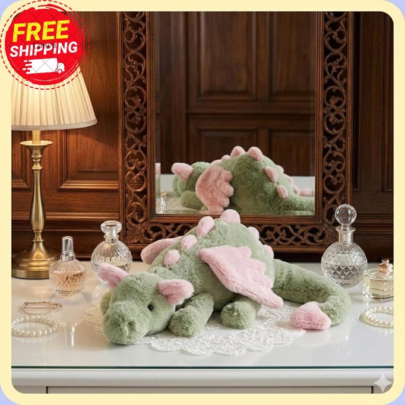 Op de afbeelding: Een groene pluche draak met roze vleugels en accenten ligt op een wit oppervlak. De weerspiegeling van de draak is te zien in een spiegel erachter. De sc&egrave;ne omvat een lamp, parfumflesjes en sieraden.