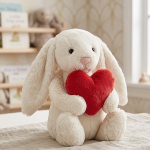 May include: A plush, cream-colored bunny rabbit holding a red heart. The bunny has long, floppy ears, a pink nose, and black eyes. The heart is a vibrant red and appears soft. The toy is sitting on a neutral-colored surface.