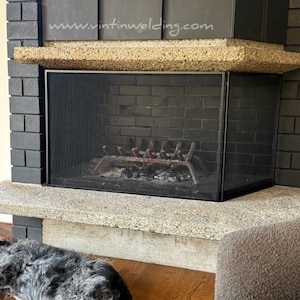 May include: A black metal fireplace screen with a fine mesh design, positioned in front of a fireplace with a textured stone hearth and mantel. The text www.vintinwelding.com is visible on the metal hood above the fireplace opening.