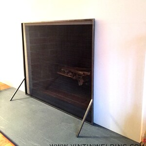 May include: A black metal fireplace screen with a mesh insert and angled legs. The screen is in front of a fireplace with logs inside. The text "www.vintinwelding.com" is visible at the bottom of the image.