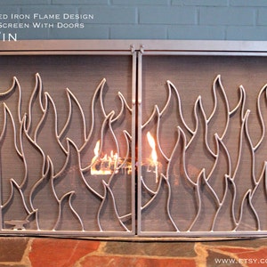 May include: Hand-forged iron fireplace screen with doors featuring a flame design. The screen is made of metal and has a black finish. The screen is in front of a fireplace with a fire burning inside. The screen is made of metal and has a black finish. The screen is in front of a fireplace with a fire burning inside.