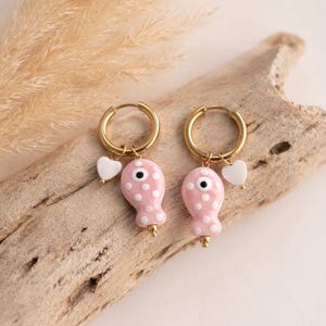 Könnte beinhalten: Goldene Creolen-Ohrringe mit rosa, gepunkteten Fisch-Anhängern. Jeder Anhänger hat ein weißes Auge und ein kleines weißes Herz. Die Ohrringe werden auf einem Stück Treibholz präsentiert.