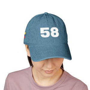May include: A blue denim baseball cap featuring the white number 58 printed on the front centre. The side of the cap displays a small rectangular flag with horizontal yellow, blue, and red stripes. The cap has a curved brim and stitched panels.