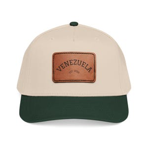 Puede incluir: Gorra de béisbol con corona color crema y visera verde oscuro. Un parche de cuero marrón en la parte delantera muestra la palabra "VENEZUELA" y "EST. 2026". La gorra tiene un botón verde oscuro en la parte superior.