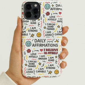 May include: White phone case with a colorful design featuring the words "Daily Affirmations" and positive affirmations like "I am strong" and "I am loved." The case has a repeating pattern of smiley faces, flowers, and stars.