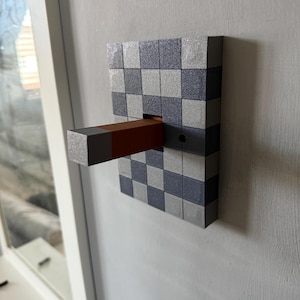 May include: A geometric wall-mounted key holder with a checkerboard pattern in grey and silver. A rectangular key holder extends outwards, featuring a brown and grey colour scheme. The key holder is mounted on a light grey wall.