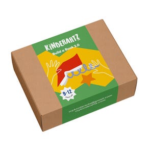 May include: A brown cardboard box with a green and yellow label. The label reads "Kinderartz Build a Book 2.0" with illustrations of hands, a book, and a star. The box is designed for ages 9-12.