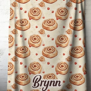 May include: A beige towel with a repeating pattern of watercolor cinnamon rolls and small red berries. The word "Brynn" is written in a cursive font at the bottom of the towel. The towel is hanging on a wooden rod.