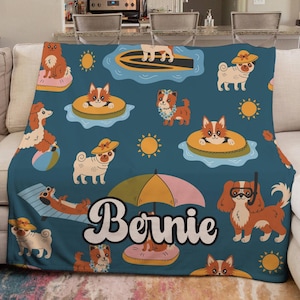 May include: A dark blue fleece blanket featuring a repeating pattern of various dog breeds engaged in summer activities like swimming, lounging on floats, and wearing sun hats, with the name Bernie printed in large white script in the center.