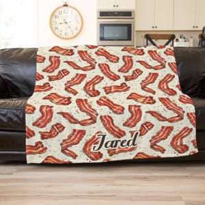 May include: A cream-colored blanket featuring a repeating pattern of cooked bacon strips in shades of red and brown. The name "Jared" is printed in a cursive font on the blanket. The blanket is draped over a black leather couch.