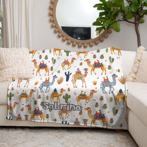 May include: A white throw blanket featuring a repeating pattern of camels, cacti, and other desert-themed elements. The camels are in various colors, and the blanket has the name "Sabrina" printed on it. The blanket is draped over a white sofa.