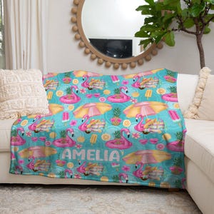 May include: A turquoise blanket with a repeating pattern of pink flamingos, yellow umbrellas, pineapples, and ice cream. The name "AMELIA" is printed in white script. The blanket is draped over a white couch.