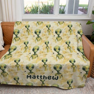 May include: A soft blanket featuring a repeating pattern of green alien figures on a cream and yellow background. The name "Matthew" is printed in dark blue at the bottom. The blanket is draped over a brown leather couch.