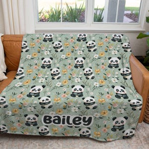 May include: A light green blanket featuring a pattern of pandas, yellow and white flowers, and green leaves. The name "Bailey" is printed in a white, rounded font. The blanket is draped over a brown leather couch.