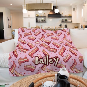 May include: A pink blanket with a pattern of cartoon bacon strips and small red hearts. The name "Bailey" is displayed in a brown font. The blanket is draped over a white couch.