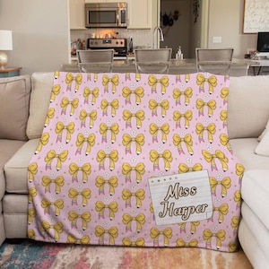 May include: A pink throw blanket with a repeating pattern of yellow bows and white flowers. A notepad with the text "Miss Harper" is attached to the blanket. The blanket is draped over a sofa.