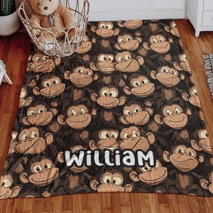 May include: A brown and tan blanket featuring a repeating pattern of smiling monkey faces. The name "William" is displayed in white block letters. The blanket is made of soft material and is suitable for a child's room.