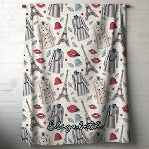 May include: A cream-colored blanket with a repeating pattern of Parisian-themed illustrations. The design includes the Eiffel Tower, striped dresses, trench coats, red hats, blue hats, red lips, and leaves. The name "Elizabeth" is written in cursive at the bottom.