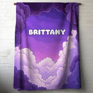 May include: A purple and white blanket with the name "BRITTANY" in white, puffy letters. The blanket features a cloud design with shades of purple and white, creating a dreamy aesthetic.