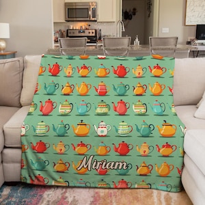 May include: A teal throw blanket featuring a pattern of colorful teapots in various designs. The blanket has a personalized name, "Miriam," printed in a cursive font. The teapots are red, yellow, orange, and blue.