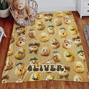 May include: A tan blanket featuring a repeating pattern of cartoon potatoes with various expressions, some wearing hats or glasses. The name "OLIVER" is displayed in a brown, bubble-style font.