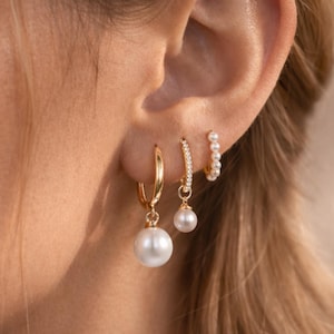 Könnte beinhalten: Nahaufnahme eines Ohrs, das mit drei Sätzen goldener Ohrringe geschmückt ist. Die Ohrringe umfassen goldene Creolen mit großen weißen Perlen, ein Paar mit kleinen Perlen und kleine Creolen mit winzigen weißen Perlen. Schmuck.