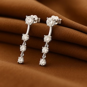 May include: A pair of silver earrings with a dangling design. Each earring features a large, round, clear gemstone at the top, followed by three smaller gemstones. The earrings are set against a brown background.