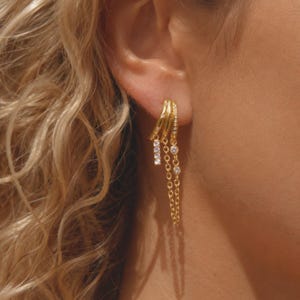 May include: A gold-toned multi-hoop earring featuring three connected bands. One band is adorned with small clear stones, while two delicate gold chains hang from the hoops, each accented with small, round, clear, sparkling stones.