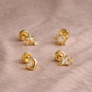 May include: Gold stud earrings with celestial motifs. The set includes a star, a sun, and a crescent moon, each with a small, clear gemstone. The earrings are presented on a pale pink surface.