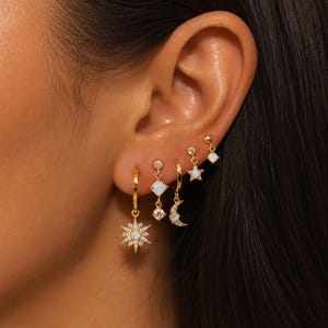 May include: A set of five gold-tone earrings featuring celestial and geometric motifs. The collection includes a starburst hoop, a square opal drop, a crescent moon charm, a small star pendant, and a simple opal stud, all accented with clear stones.