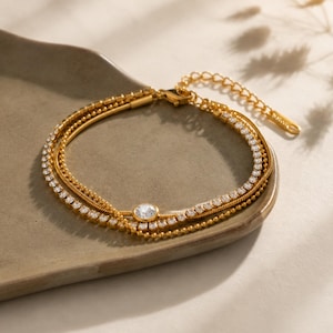 Puede incluir: Una pulsera dorada de varias capas que presenta una cadena fina, una cadena de cuentas y una cadena tipo tenis con piedras transparentes. Una piedra redonda transparente está engastada en el centro. Incluye una cadena de extensión ajustable.