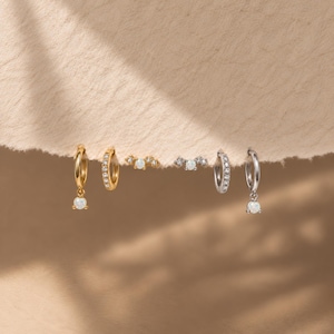 May include: A collection of six earrings including two gold hoop earrings with dangling white stones, one gold hoop earring with embedded stones, one gold stud earring with three stones, one silver stud earring, and two silver hoop earrings with stones.