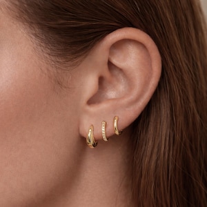 May include: Three gold hoop earrings of varying sizes are displayed on an ear. The largest hoop has a smooth, rounded design. The middle hoop is adorned with small, sparkling stones. The smallest hoop has a simple, curved shape.