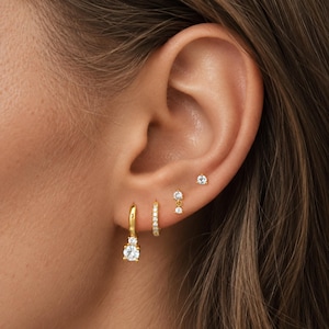 Könnte beinhalten: Ein Set aus vier goldfarbenen Ohrringen mit klaren, rund geschliffenen Steinen. Das Set besteht aus einem Creolen-Ohrring mit hängendem Stein, einer kleinen Pavé-Creole, einem Ohrstecker mit zwei Steinen und einem mit einem Stein.