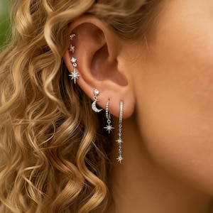 Könnte beinhalten: Eine Auswahl an silbernen Ohrringen mit Himmelsmotiven. Die Ohrringe zeigen Stern- und Halbmond-Designs, einige verziert mit winzigen, funkelnden Steinen. Die Ohrringe werden an einem Ohr getragen, mit lockigem blondem Haar im Hintergrund.