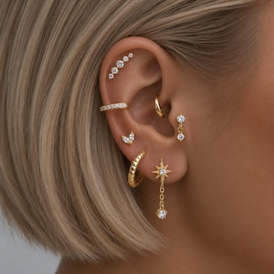 May include: A collection of gold-toned earrings featuring a curved bar with five clear stones, a plain hoop, a marquise-cut stone stud, a small hoop, a dangling two-stone earring, and a starburst charm earring with a dangling clear stone.