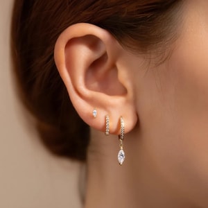 May include: Close-up of a person's ear adorned with three gold earrings. The smallest is a stud, the middle one is a small hoop, and the largest is a hoop with a dangling, marquise-shaped charm, all encrusted with small, clear stones.