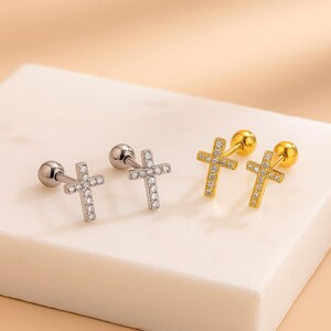 May include: Two pairs of cross-shaped stud earrings. One pair is silver-toned with clear stones, and the other is gold-toned with clear stones. The earrings are displayed on a white surface.