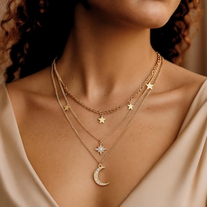 Puede incluir: Un conjunto de collares dorados en capas con un colgante de media luna con piedras transparentes, un colgante de estrella y múltiples dijes de estrella. Los collares se muestran en el cuello de una persona, con un fondo neutro.