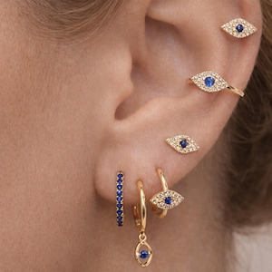 Puede incluir: Primer plano de una oreja adornada con pendientes de oro con detalles de zafiro azul. Los pendientes incluyen pequeños aros, tachuelas en forma de ojo y un dije colgante. La joyería se encuentra sobre el fondo del tono de piel natural de la oreja.